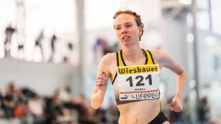 Eine Athletin mit der Nummer 121 auf ihrem Trikot läuft in einem Rennen, wahrscheinlich in einer Halle. Sie trägt ein gelbes und schwarzes Trikot mit einer Halskette und einem Armband.