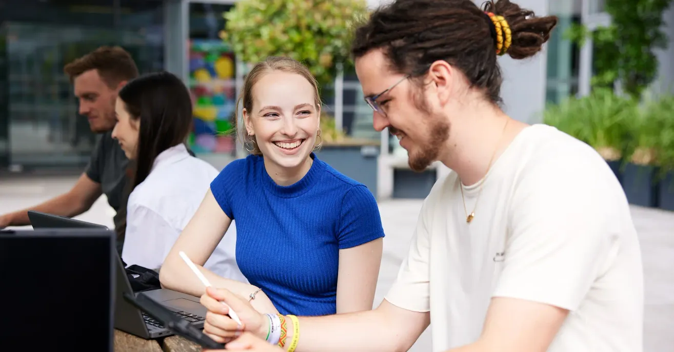 Eine Frau in einem blauen Shirt lächelt, während sie einen Stift hält, neben einem Mann mit Brille. Sie sitzen an einem Tisch, wahrscheinlich in einer ungezwungenen Umgebung.