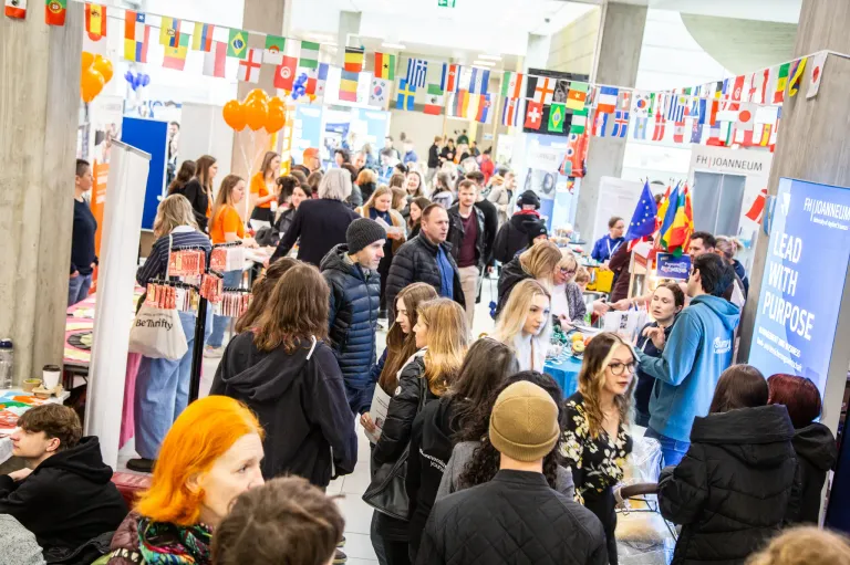 Eine belebte Indoor-Veranstaltung mit vielen Menschen in verschiedenen Kleidungsstilen, möglicherweise eine Messe mit Ständen und Verkaufsbuden. Flaggen verschiedener Länder hängen darüber.