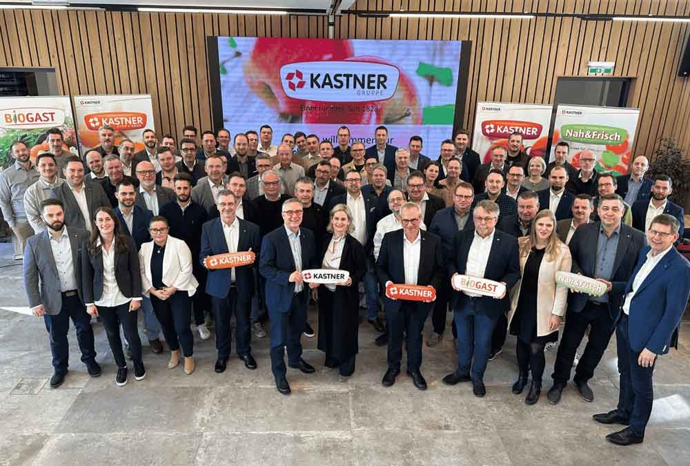 A group of people are posing for a photo, holding signs that say Kastner. They are wearing formal attire. Behind them is a large screen with the Kastner logo and the text 'Einer für alle'. There are banners with the Kastner logo and the text 'Nah & Frisch'.