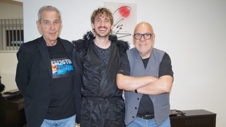 Drei Männer stehen zusammen in einem Studio, einer lächelt und die anderen haben die Arme verschränkt. Der Mann in der Mitte trägt einen schwarzen Umhang. Hinter ihnen befindet sich eine Wand mit einem abstrakten Gemälde.