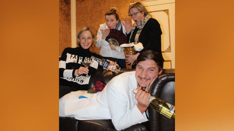 Eine Gruppe von Menschen posiert für ein Foto, zwei Frauen halten Weingläser und eine Flasche Schnaps, ein Mann hält eine Rose und eine andere Frau eine Fächer.