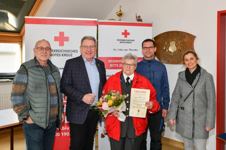 Five people stand close together in front of a wall with a Red Cross banner. A woman in a red jacket holds a bouquet and a certificate. The men wear glasses, suits, and jackets. Behind them, there is a decorative wooden plaque with a golden design.