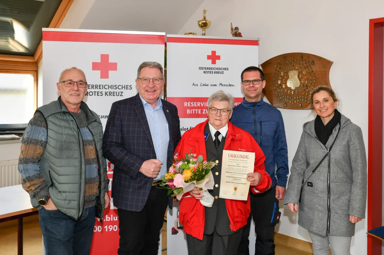 Five people stand close together in front of a wall with a Red Cross banner. A woman in a red jacket holds a bouquet and a certificate. The men wear glasses, suits, and jackets. Behind them, there is a decorative wooden plaque with a golden design.