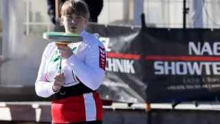 Eine junge Frau hält einen Diskus und bereitet sich darauf vor, ihn bei einer Outdoor-Sportveranstaltung zu werfen. Dahinter steht eine verschwommene Figur in der Nähe eines Banners, das 'Technik' besagt.