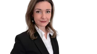 Eine Frau in einem schwarzen Blazer und weißem Hemd, lächelt und posiert für ein Foto vor schwarzem Hintergrund.