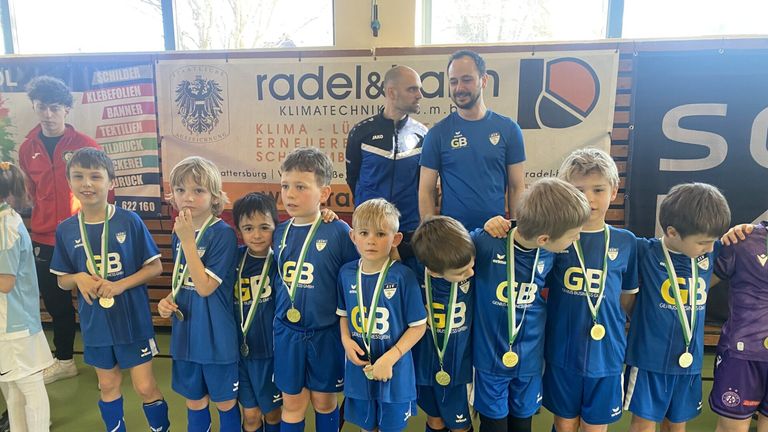 Eine junge Fußballmannschaft in blauen Trikots und mit Medaillen posiert für ein Foto mit zwei Trainern. Sie stehen vor einem Banner mit Logos und Text.