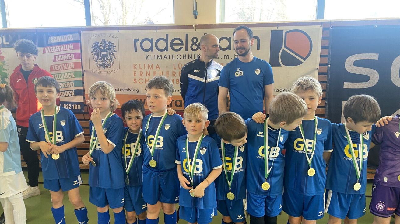 Eine junge Fußballmannschaft in blauen Trikots und mit Medaillen posiert für ein Foto mit zwei Trainern. Sie stehen vor einem Banner mit Logos und Text.