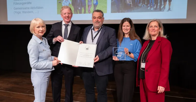 Five people stand on stage with certificates. The man in the middle holds a certificate, while the two women hold glass awards. The man on the left smiles, and the man on the right has a beard. Behind them is a projector screen.