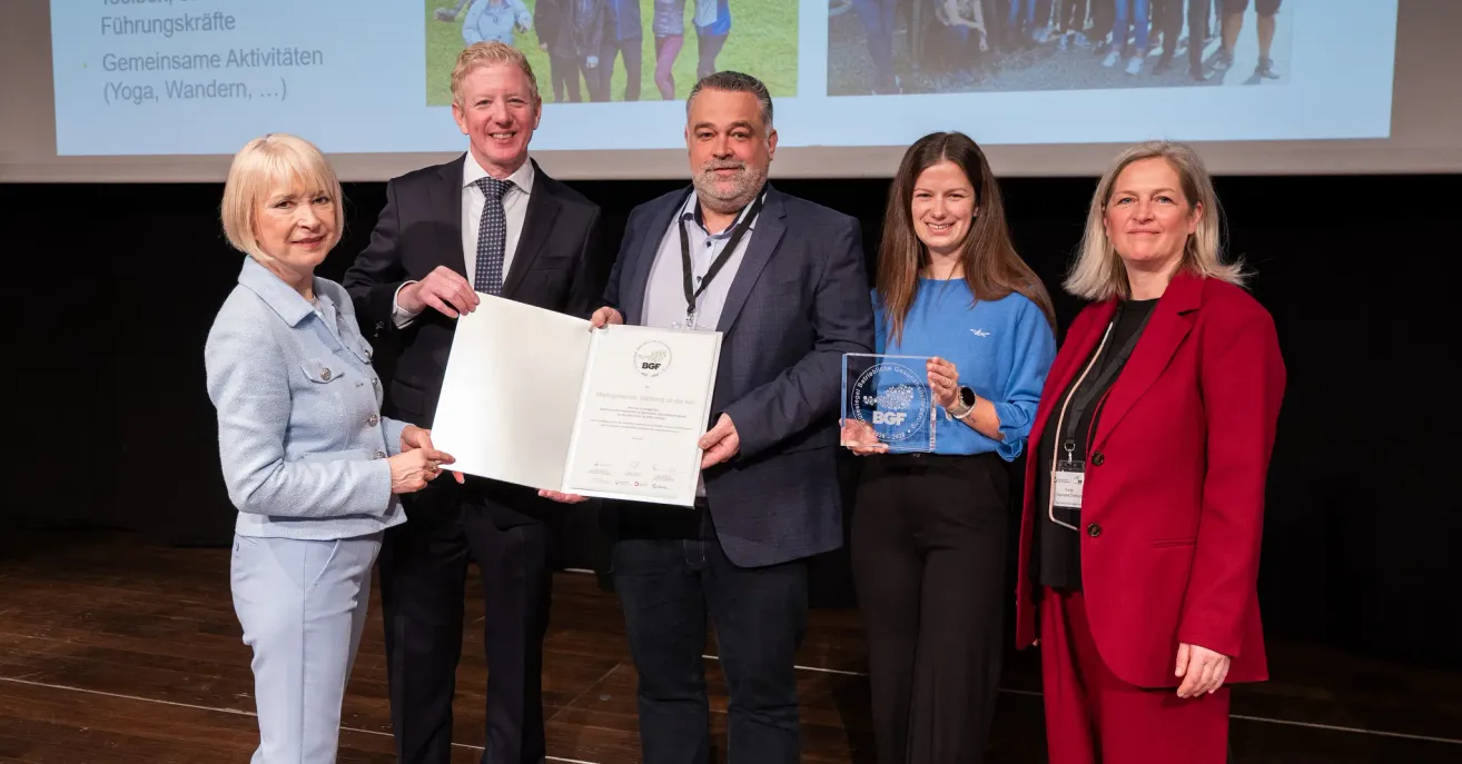 Five people stand on stage with certificates. The man in the middle holds a certificate, while the two women hold glass awards. The man on the left smiles, and the man on the right has a beard. Behind them is a projector screen.