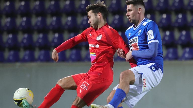 Zwei Fußballspieler in Trikots kämpfen auf einem Feld. Einer in Rot, der andere in Blau. Der Spieler im roten Trikot versucht, den Ball zu stehlen.