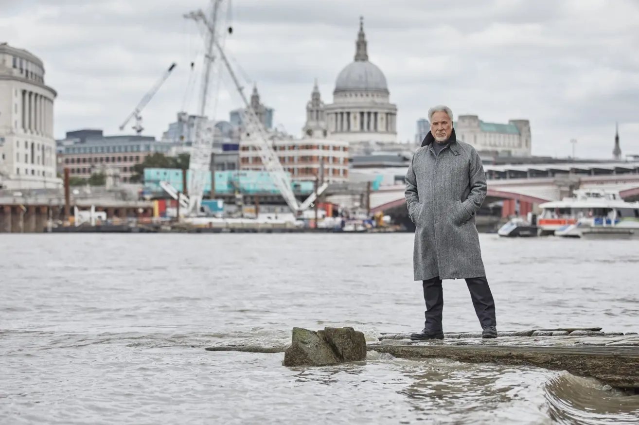 Ein älterer Mann steht auf einem Felsen im Fluss, trägt einen grauen Mantel und blickt auf die Skyline der Stadt London.