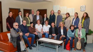 Eine Gruppe von Menschen posiert für ein Foto in einem Raum mit Sofas und einem Tisch, wahrscheinlich bei einer Veranstaltung. Einige tragen Brillen und halten Gegenstände wie einen Stock und eine Handtasche.