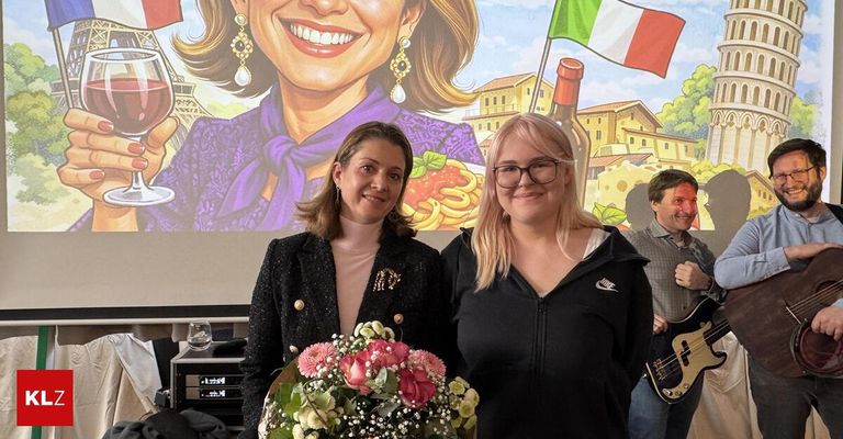 Zwei Frauen stehen nebeneinander und halten einen Blumenstrauß. Dahinter ist ein großes Poster einer lächelnden Frau und ein Mann mit einer Gitarre.