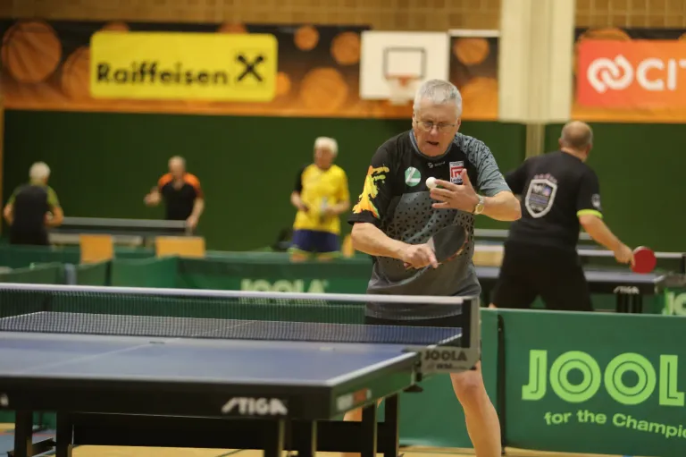 Ein älterer Mann spielt Tischtennis in einer Halle, hält einen Ball und einen Schläger. Im Hintergrund spielen zwei andere. Ein Banner zeigt "Joola" und "Raiffeisen".