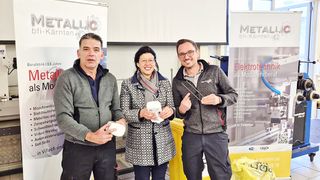 Drei Personen stehen nebeneinander, halten Tassen und lächeln, in einem Raum mit einer weißen Wand und zwei Bannern. Die Banner haben die Worte 'METALLIC' und 'bfi-Kärnten'. Sie sind wahrscheinlich bei einer Veranstaltung.
