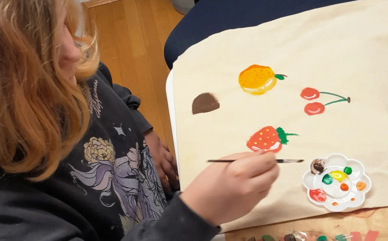 Eine Person malt auf einer Leinwand mit Früchten und Blumen. Die Leinwand steht auf einem Tisch. Die Person trägt ein schwarzes Langarmshirt.