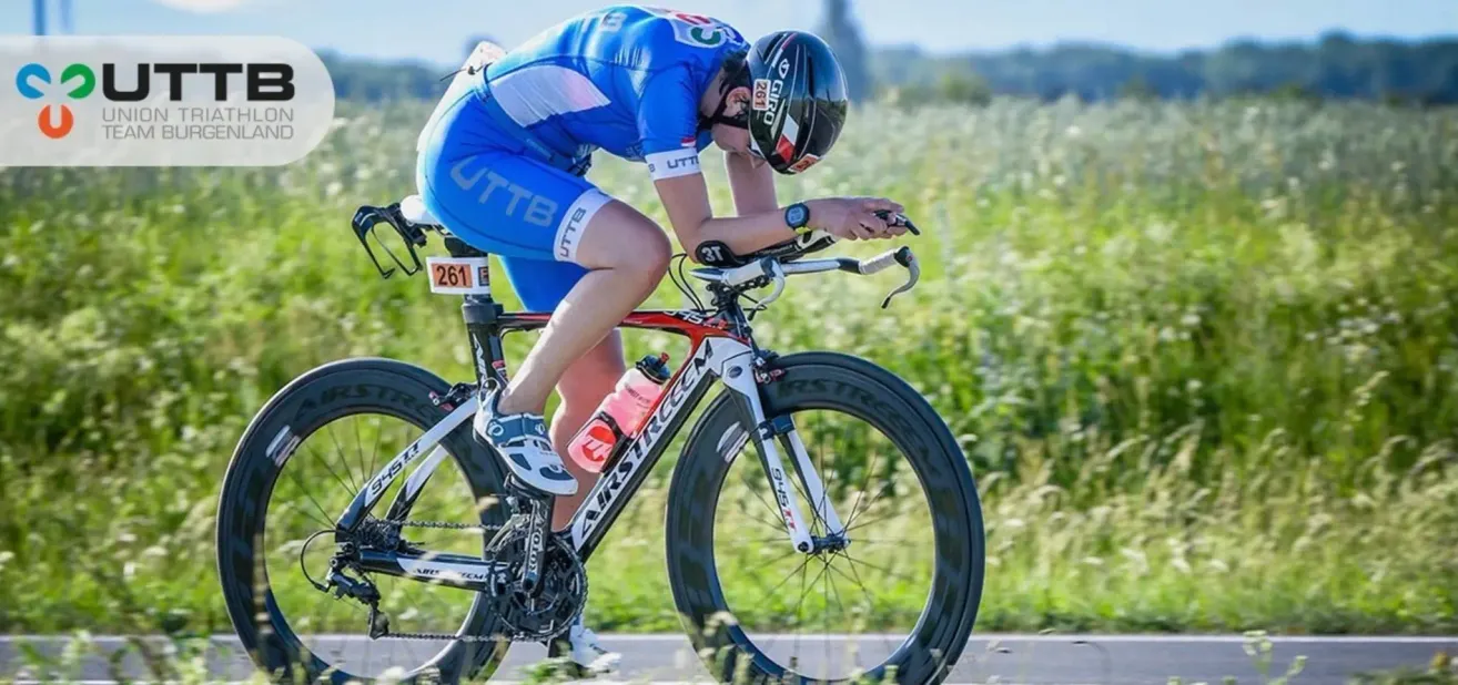 Ein Radfahrer in einem blauen Rennanzug beugt sich vor, während er ein Fahrrad auf einer grasigen Straße fährt. Eine Flasche ist am Fahrrad befestigt. Die Nummer 261 befindet sich auf dem Fahrrad.