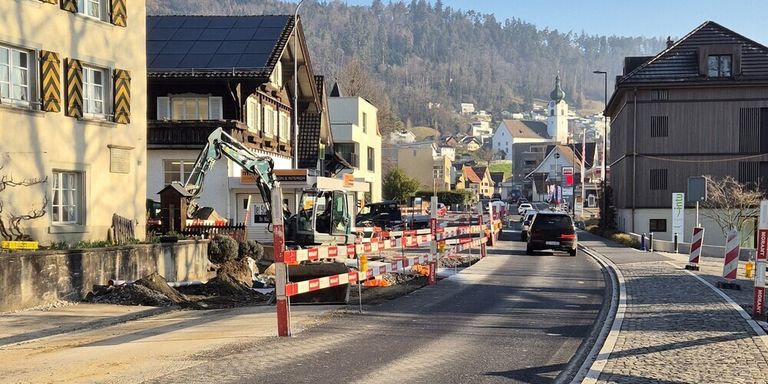 A street under construction with barricades and machinery, with a car driving down the road. Buildings, trees, and mountains in the background.