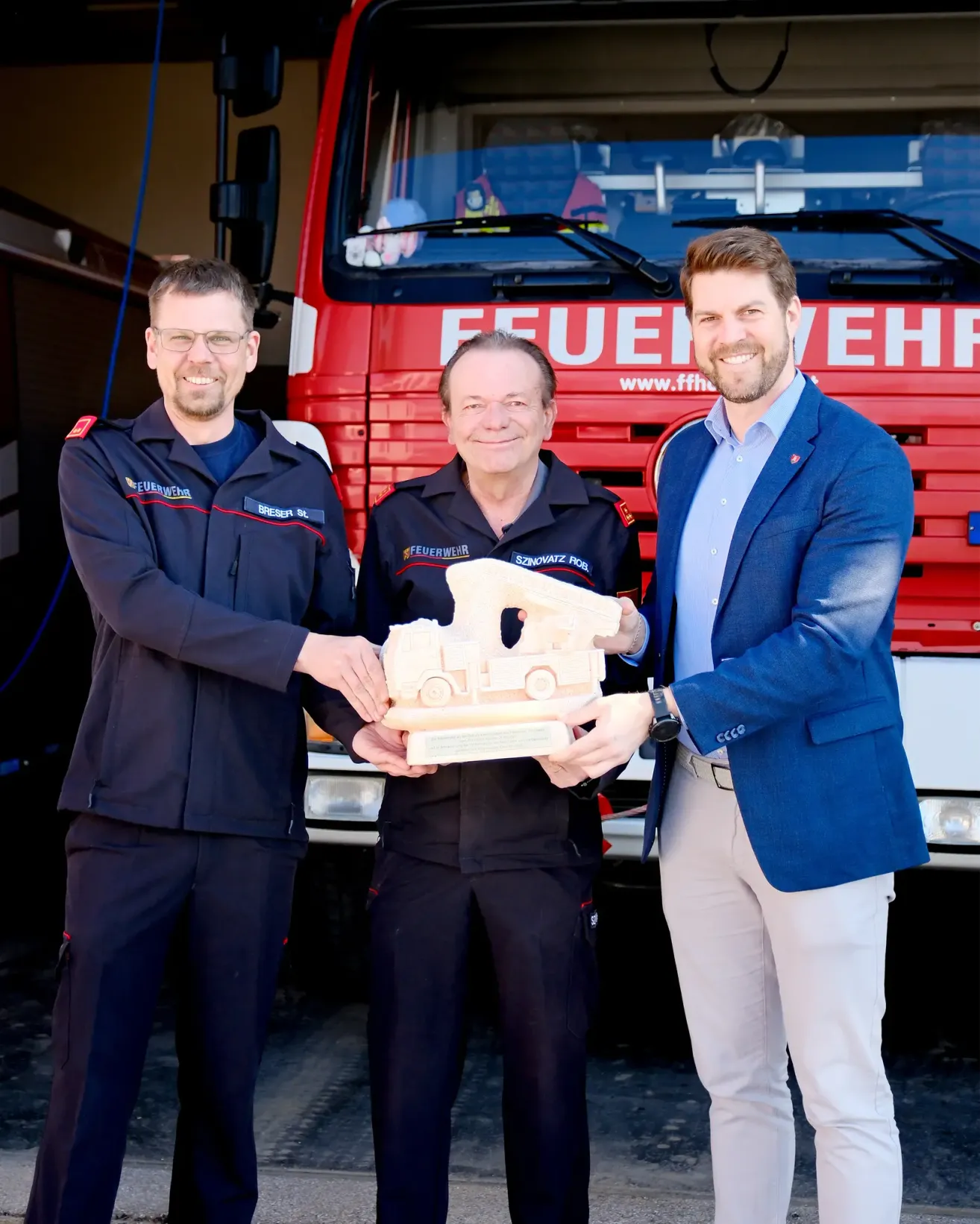 Drei Männer stehen vor einem roten Feuerwehrauto, zwei in blauen Uniformen, einer in einem blauen Anzug, lächelnd, halten ein Holzspielzeug-Feuerwehrauto.