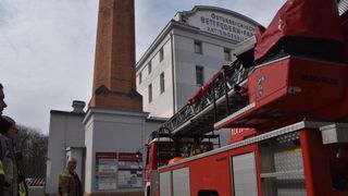 Ein Feuerwehrauto mit ausgefahrener Leiter steht vor einem großen Gebäude mit einem hohen Schornstein aus Ziegeln. Ein Feuerwehrmann steht in der Nähe.