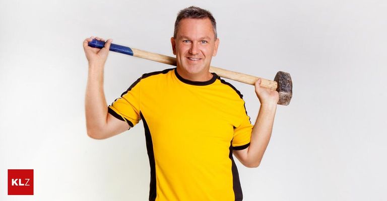 Ein Mann in einem gelben und schwarzen T-Shirt hält einen Hammer über dem Kopf und lächelt.