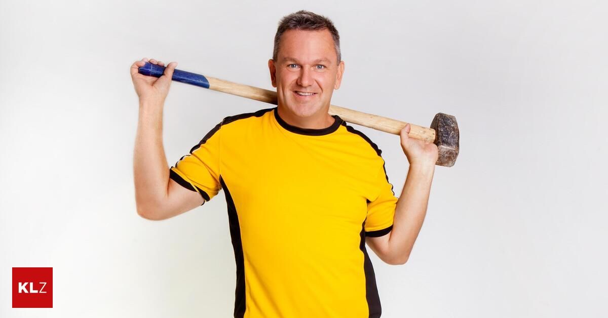 Ein Mann in einem gelben und schwarzen T-Shirt hält einen Hammer über dem Kopf und lächelt.