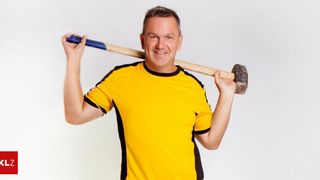 Ein Mann in einem gelben und schwarzen T-Shirt hält einen Hammer über dem Kopf und lächelt.