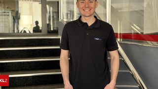 Ein Mann steht in der Nähe einer Treppe und trägt ein schwarzes Poloshirt mit Logo. Er lächelt und posiert für ein Foto. Hinter ihm ist eine Glaswand mit einer Reflexion.