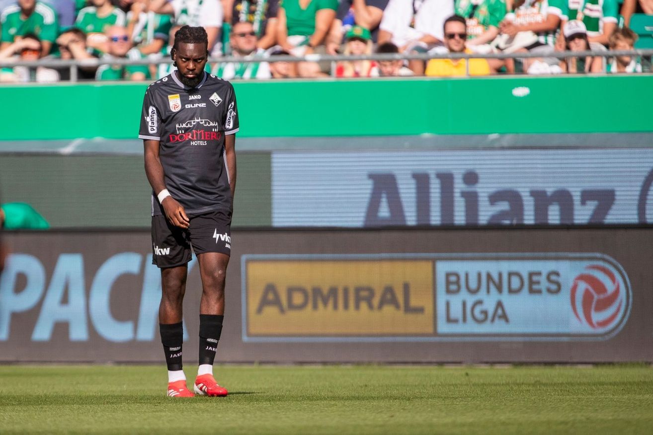 Ein Fußballspieler in einem schwarzen Trikot steht auf dem Feld und schaut nach unten. Hinter ihm zeigt ein grüner Banner 'Allianz' und 'Admiral Bundesliga'. Zuschauer in grünen und weißen Trikots beobachten von den Tribünen aus.