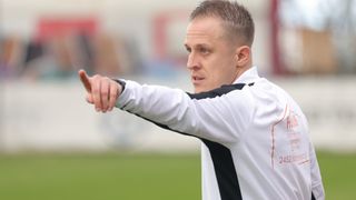 Ein Mann in einem weißen und schwarzen Langarmshirt zeigt mit dem Finger nach links, vermutlich während einer Sportveranstaltung auf einem Rasenfeld.