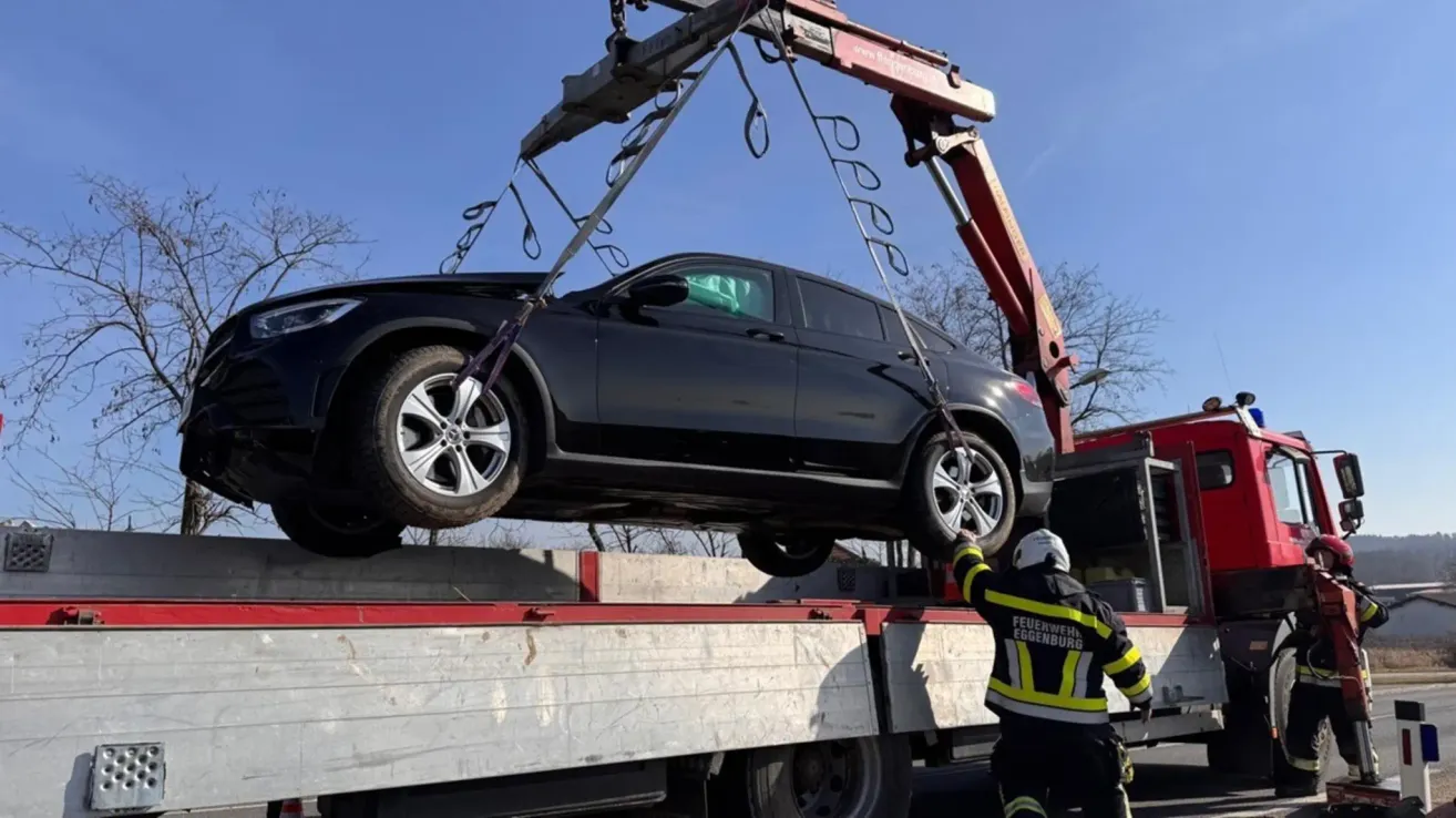 A fireman wearing a safety helmet helps lift a damaged black SUV using a crane on a flatbed truck.