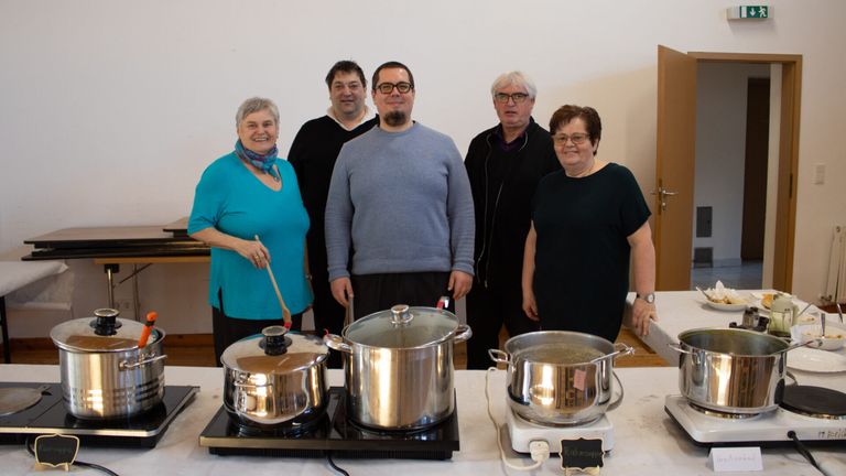 Five individuals stand behind a table with three pots. The man in the middle wears glasses and a gray sweater. The woman on the left holds a wooden spoon. The door is open.