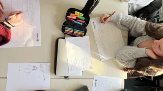 Mehrere Schüler sitzen an einem Schreibtisch und arbeiten an einem Projekt. Sie haben Papiere, Bleistifte und einen Bleistiftkasten mit verschiedenen farbigen Stiften. Die Person auf der rechten Seite hält einen Bleistift und einen Stift. Die Person auf der linken Seite hält einen Stift. Die Person in der Mitte trägt eine Brille.