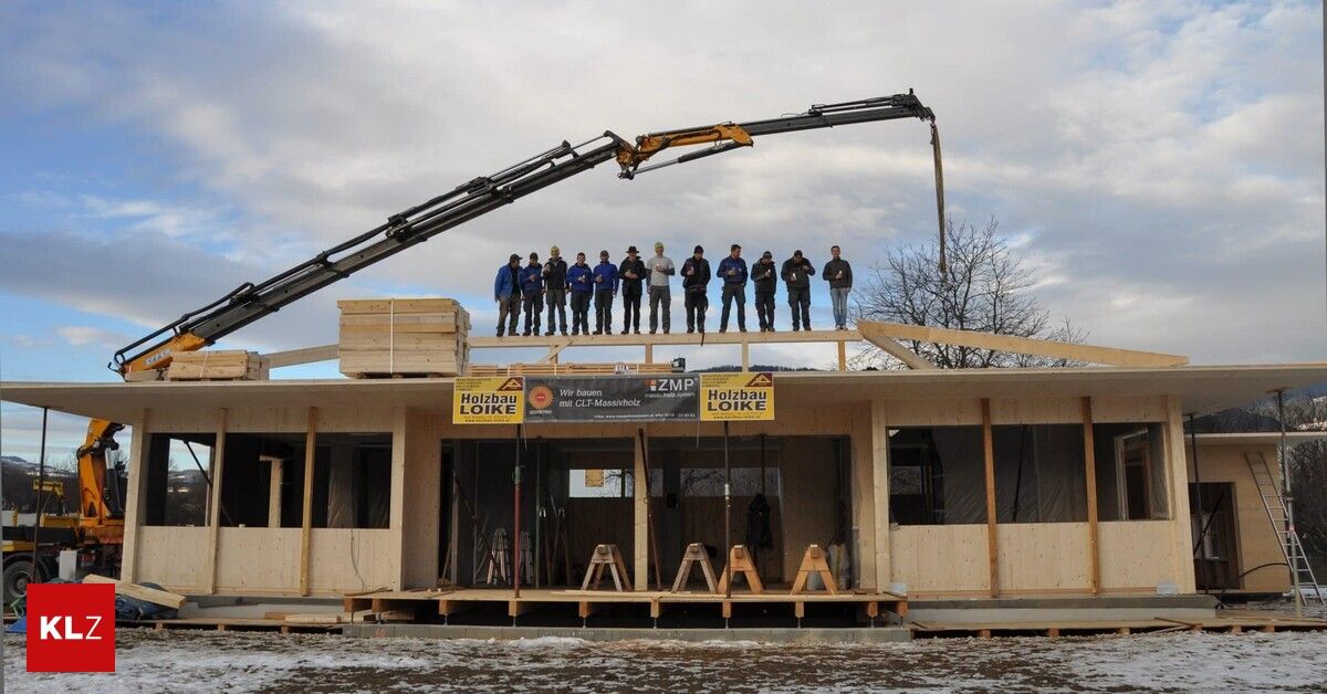 Eine Gruppe von Menschen in Winterkleidung steht auf einem im Bau befindlichen Holzhaus, wobei ein Kran einen Holzbalken über ihnen hebt. Das Haus hat ein Schild mit der Aufschrift Holzbau Loike.