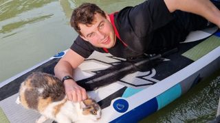 Ein Mann in einem schwarzen T-Shirt liegt lächelnd auf einem aufblasbaren Boot. Neben ihm sitzt eine Katze, die direkt in die Kamera schaut.