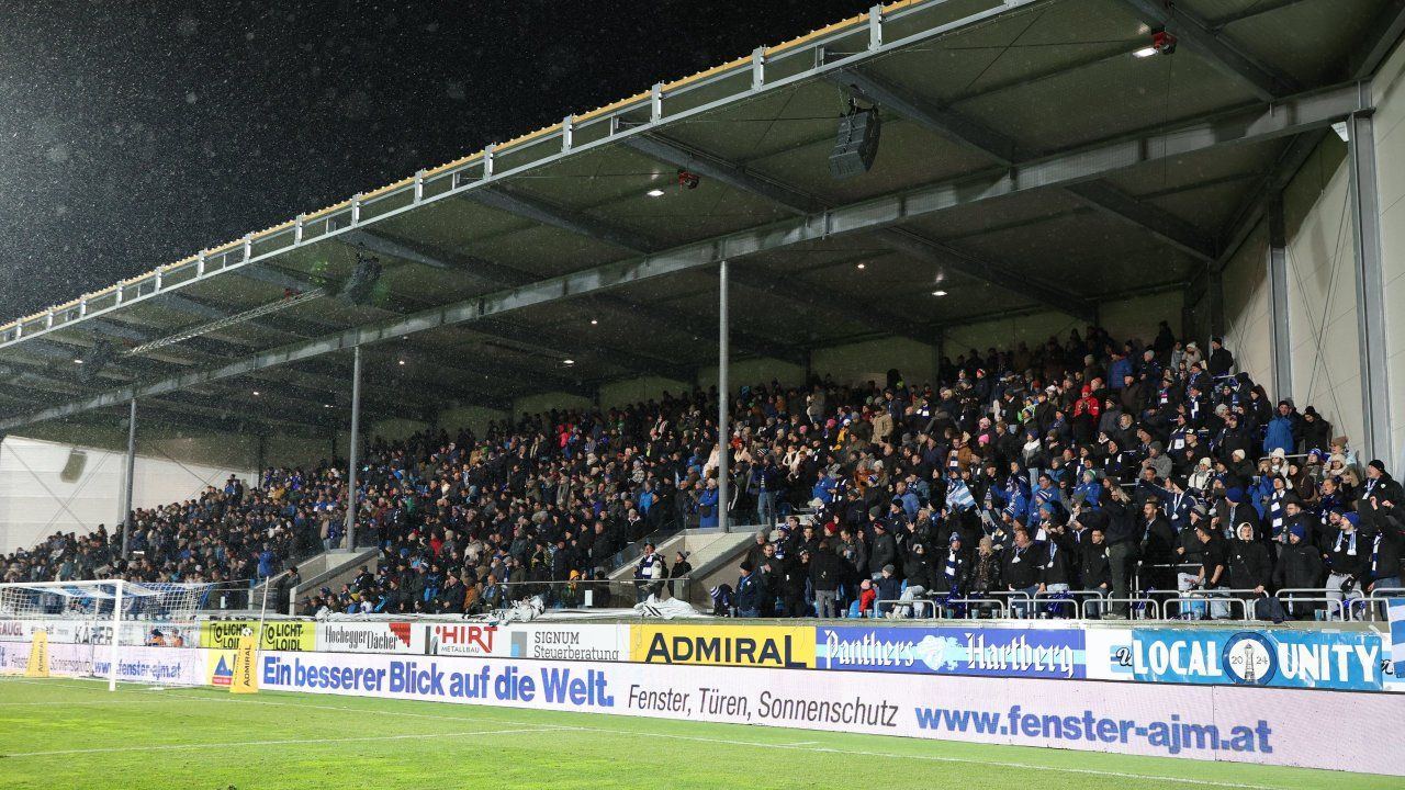 Ein Nachtstadion gefüllt mit Zuschauern, die Blau und Weiß tragen, in Reihen stehen und eine Veranstaltung auf dem Feld verfolgen.