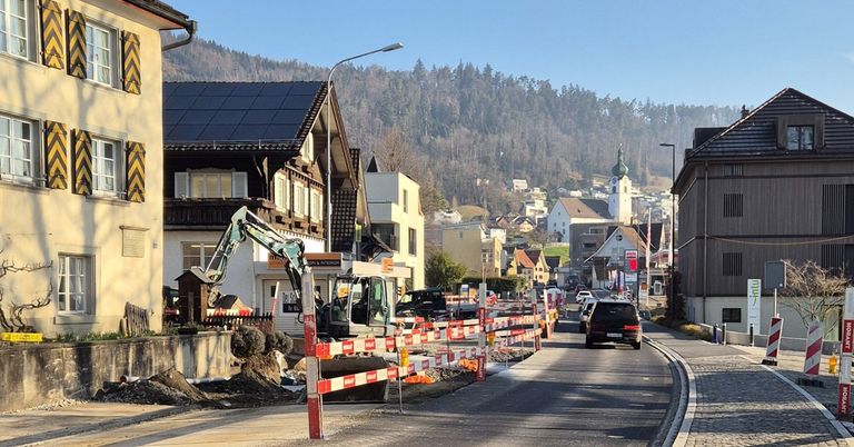 A street with construction barriers, an excavator, and houses in a mountainous area. A black car drives down the road.