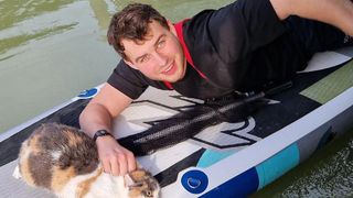 Ein lächelnder Mann liegt mit seiner Katze auf einem Paddelboard. Er trägt ein schwarzes Shirt und die Katze hat gelbe Augen.