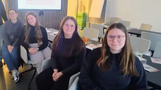 Drei Frauen sitzen in einem Klassenzimmer und schauen in die Kamera. Hinter ihnen ist eine Projektionsleinwand sichtbar, die Text anzeigt. Das Klassenzimmer ist mit Tischen, Stühlen und einer Tafel ausgestattet.