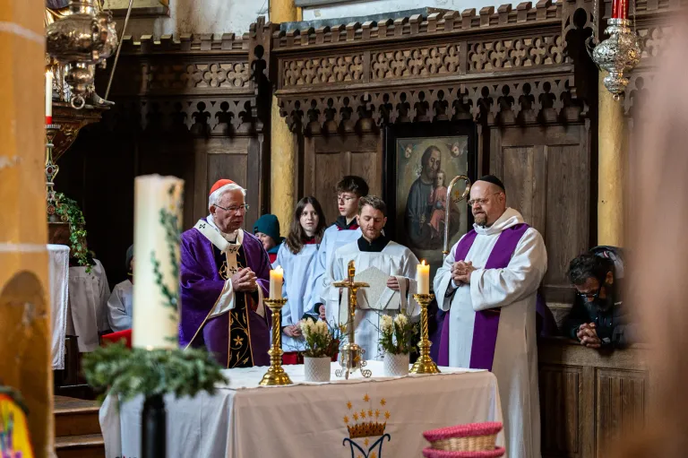 A religious ceremony is being held in a church. The priests are praying in front of the altar with candles. People are standing behind them. A wooden cross and flowers are on the altar. A painting is on the wall behind them.