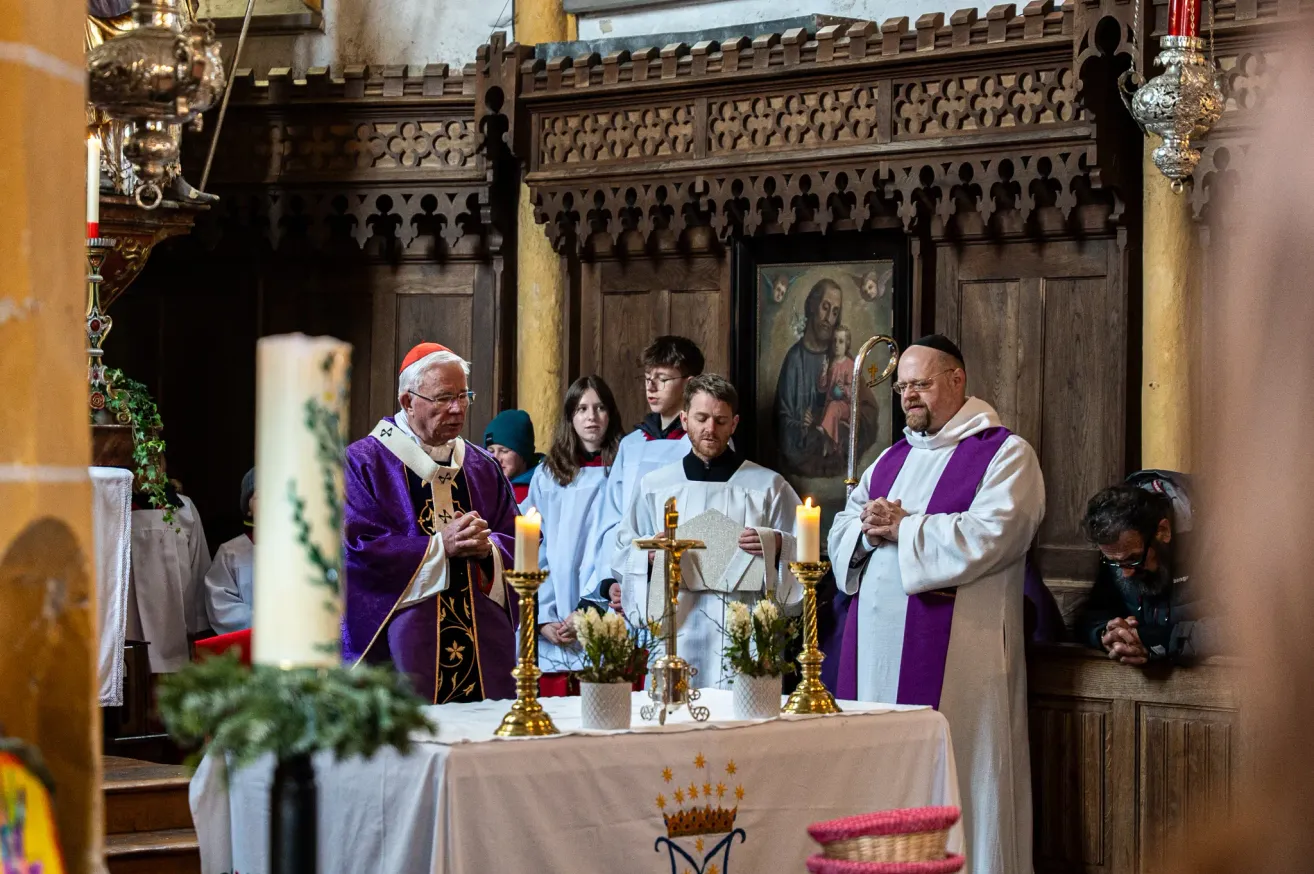 A religious ceremony is being held in a church. The priests are praying in front of the altar with candles. People are standing behind them. A wooden cross and flowers are on the altar. A painting is on the wall behind them.