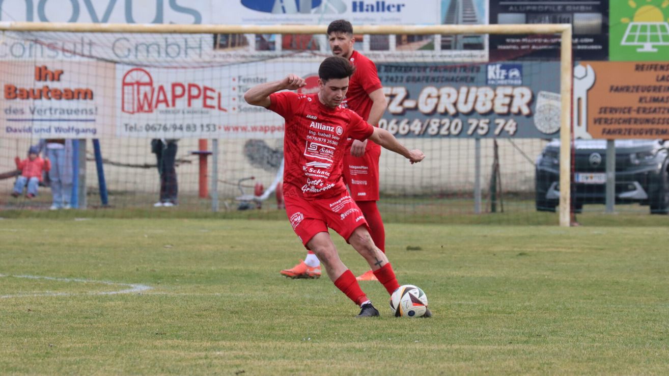 Ein Fußballspieler in einem roten Trikot tritt einen Ball auf einem Rasenfeld. Hinter ihm steht ein weiterer Spieler, und sie spielen in einem Stadion mit Zaun.