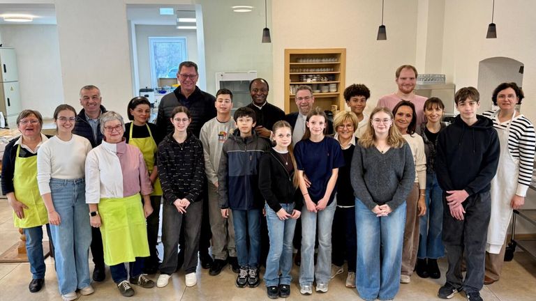 A group of people are posing for a photo in a room with a shelf of plates and cups. The individuals include children and adults, all smiling and looking at the camera. The room has white walls and a ceiling lamp.