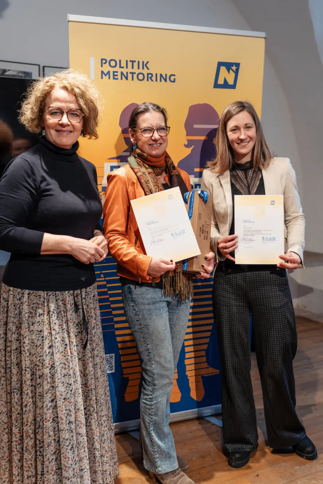 Drei Frauen in formeller Kleidung stehen vor einem farbenfrohen Hintergrund und halten Urkunden in den Händen. Die Frau auf der linken Seite trägt einen Blumenrock und eine Brille. Die Frau in der Mitte hat einen Schal und hält eine Urkunde und eine braune Schachtel. Die Frau auf der rechten Seite trägt einen Blazer und hält ebenfalls eine Urkunde.