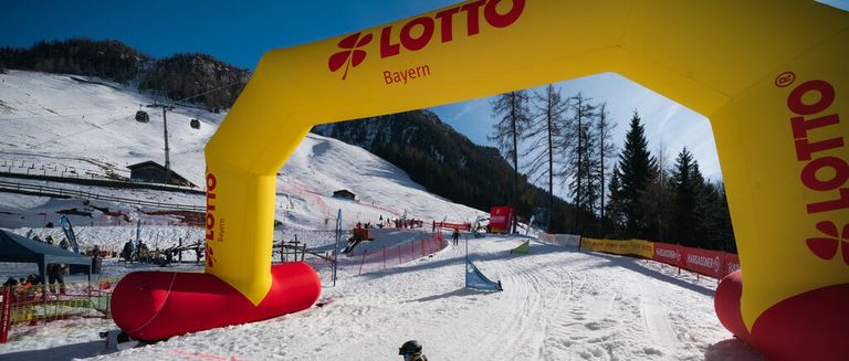Eine verschneite Skipiste mit einem gelben Bogen, der mit Lotto Bayern beschriftet ist. Mehrere Leute steigen die Piste hinunter, und einige stehen herum.