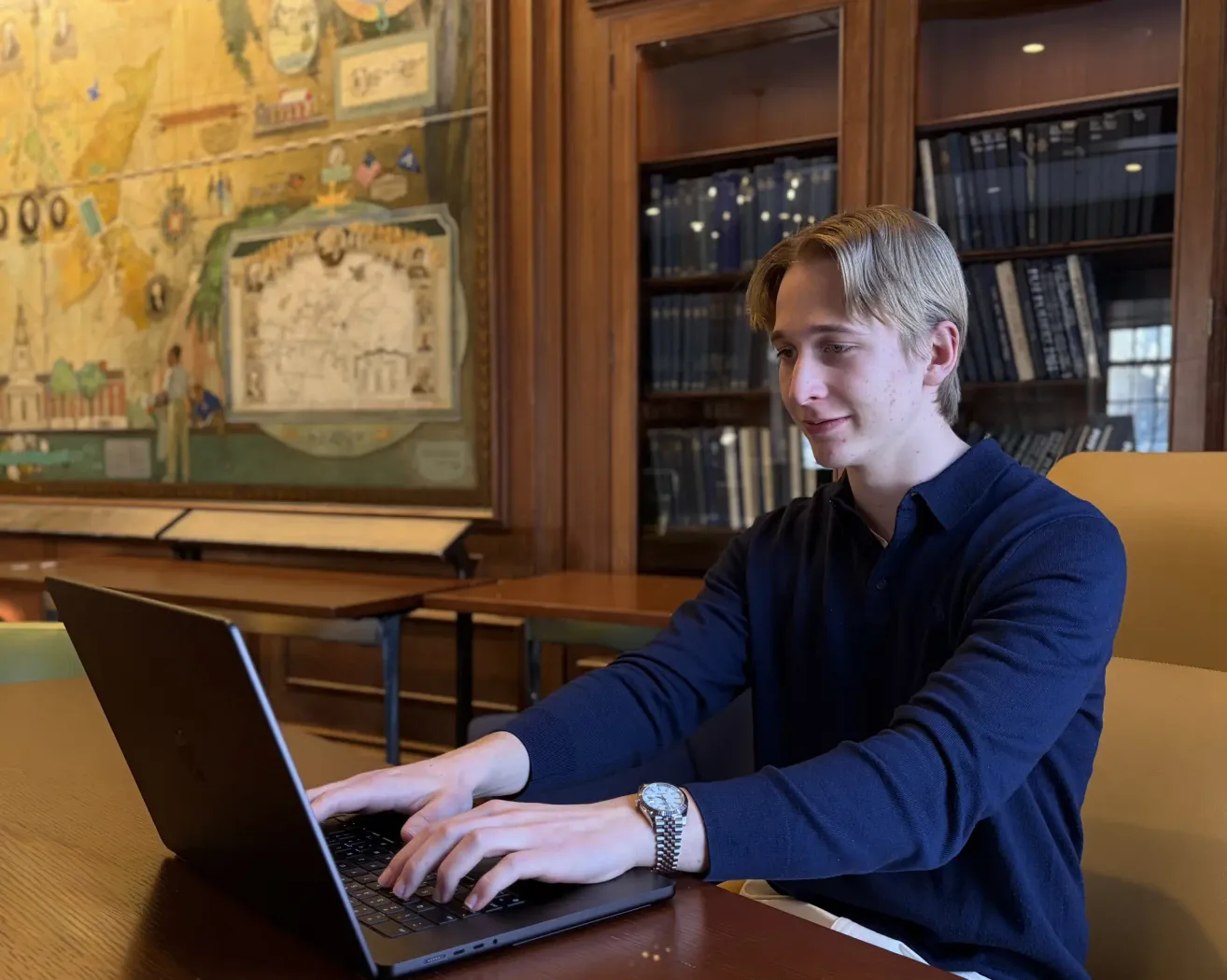 A young man in a blue long-sleeve shirt sits at a desk, working on a laptop. Behind him, a bookshelf is filled with books, and a mural decorates the wall.