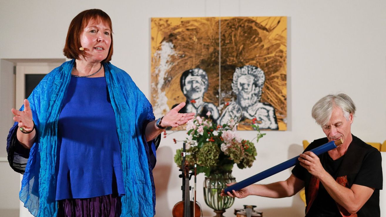 Eine Frau in einem blauen Kleid spricht und gestikuliert vor einem großen Gemälde. Neben ihr hält ein Mann ein blaues Objekt, möglicherweise ein Musikinstrument.