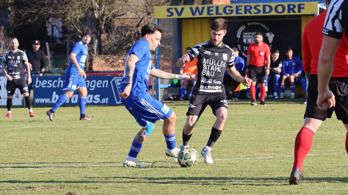 Zwei Fußballspieler, einer in Blau und der andere in Schwarz, sind in einem Fußballspiel auf einem Feld. Der Spieler in Blau versucht, den Ball zu kontrollieren, während der andere Spieler in Schwarz versucht, ihn zu stehlen.