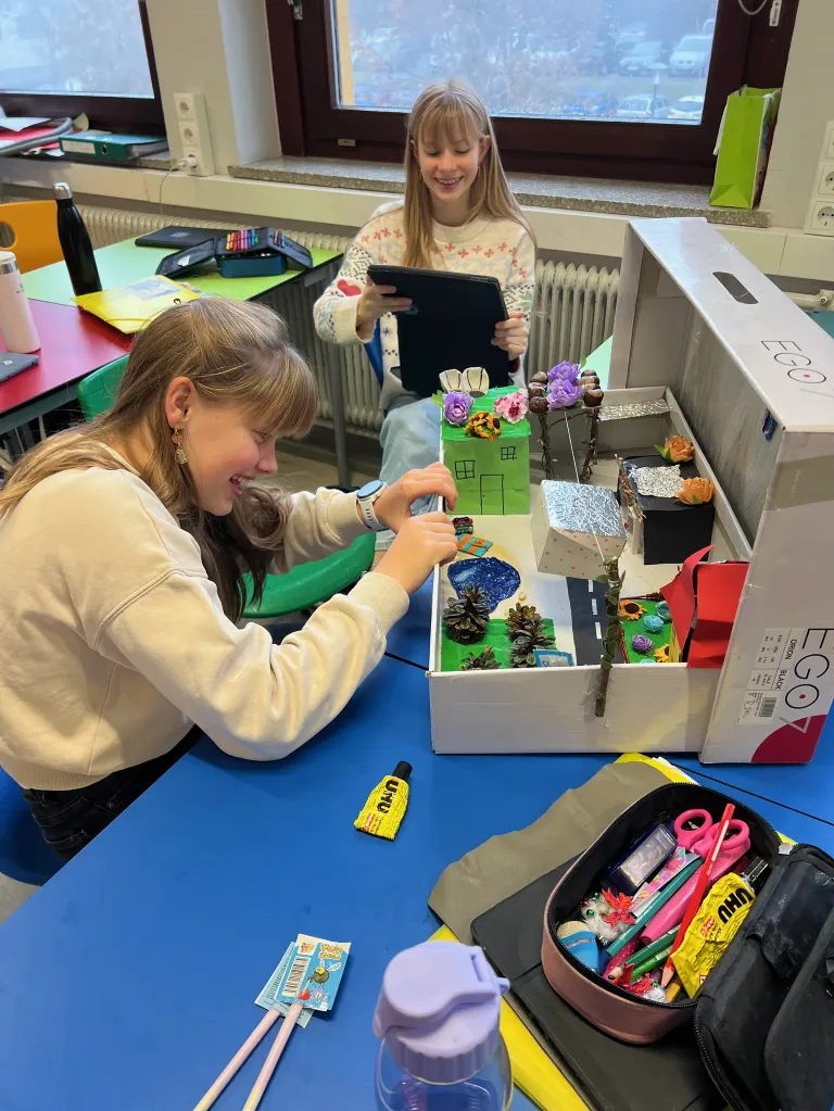 Two girls in a classroom, one using a tablet and the other decorating a small house model in a cardboard box.